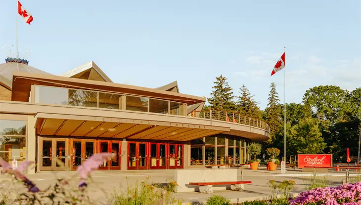  Das Stratford Festival Theatre im Sommer, mit roten Türen, wehenden kanadischen Flaggen und blühenden Gärten im Vordergrund unter einem klaren Himmel.