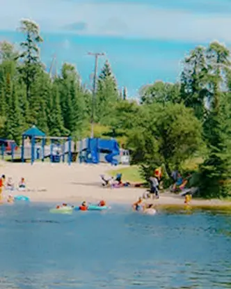 Familles profitant d’un jour d’été sur l’une des plages de sable de Kenora, avec des enfants nageant et jouant près du rivage entouré d’arbres.