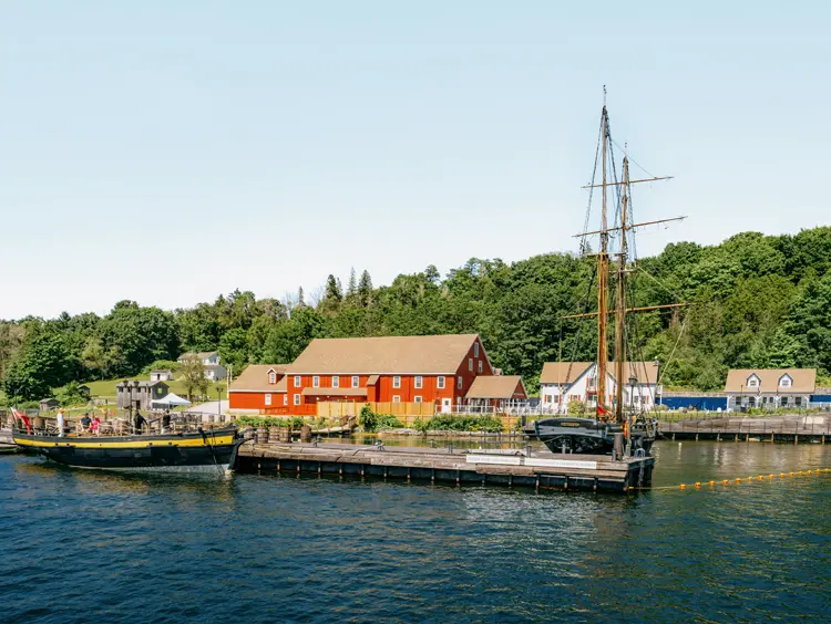 Un gran velero atracado en Discovery Harbour en Penetanguishene, Ontario, con edificios patrimoniales de color rojo y una exuberante vegetación bajo un cielo claro de verano.