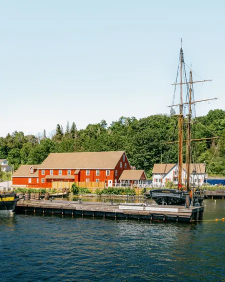 Ein Großsegler liegt im Discovery Harbour in Penetanguishene, Ontario, mit roten historischen Gebäuden und üppigem Grün unter einem klaren Sommerhimmel.