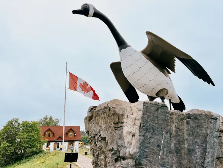 Large black and white Wawa Goose monument with wings outstretched, perched on a tall rock, with a Canadian flag and a building in the background.