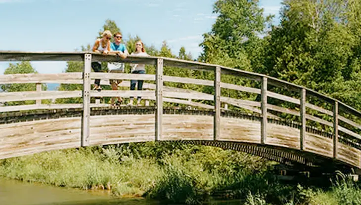  インバーヒューロン州立公園のアーチ型の木製の橋の上に立つ 3 人が、豊かな緑に囲まれた晴れた夏の日を楽しんでいます。