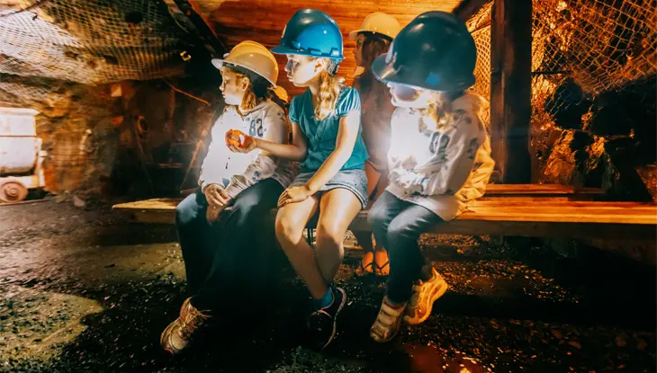 Vier Kinder mit Schutzhelmen sitzen auf einer Holzbank in einer schwach beleuchteten unterirdischen Höhle mit Felswänden bei Dynamic Earth. 