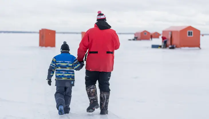 晴れた冬の日に、父と息子が凍ったシムコー湖を渡り、氷上釣り小屋に向かって歩いています。 