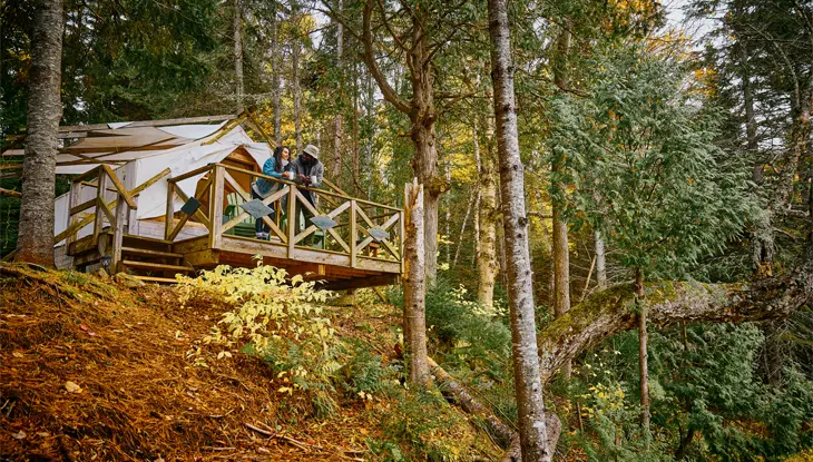 Two people standing on the deck of a wooden cabin, perched on a hill overlooking lush forest at Bartlett Lodge, Algonquin.
