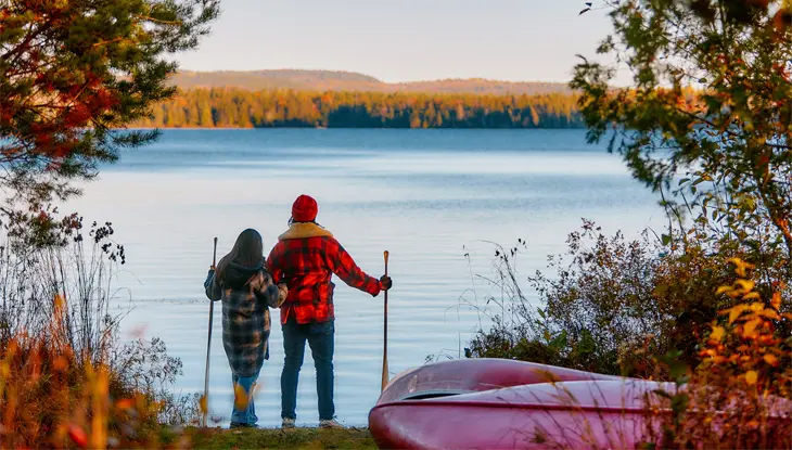 Ein Paar hält Wanderstöcke in der Hand und blickt auf einen stillen See, der von herrlichem Herbstlaub umgeben ist.