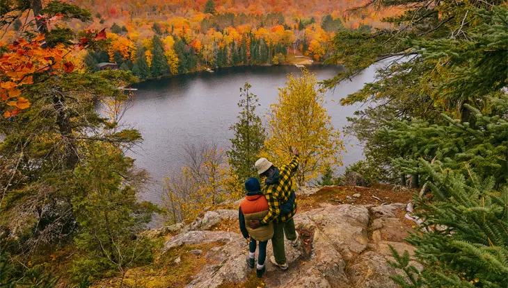 두 명의 하이커가 고요한 호수와 광활한 주황색과 붉은색 숲을 바라보는 전망대에 서 있습니다.