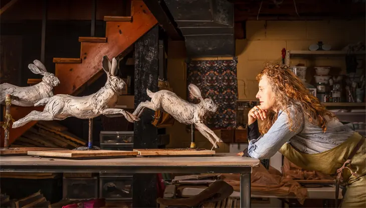 Una escultura de tres conejos saltando mira a una mujer con bata que se inclina hacia ellos pensativamente.