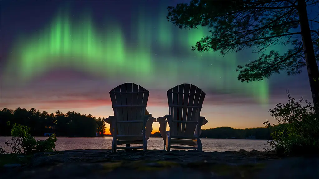 Dos sillas Adirondack dan a un tranquilo lago en Muskoka, Canadá, por la noche, frente a la colorida aurora boreal que danza en el cielo.