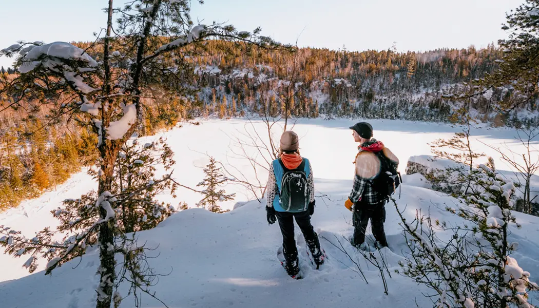 Dos personas que caminan con raquetas de nieve en el norte de Ontario se detienen para admirar la vista del bosque cubierto de nieve sobre un lago congelado.