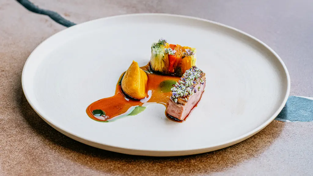 Ein elegant serviertes Gericht aus Fleisch und Gemüse mit Soße, daneben hält eine Frauenhand ein Weinglas.