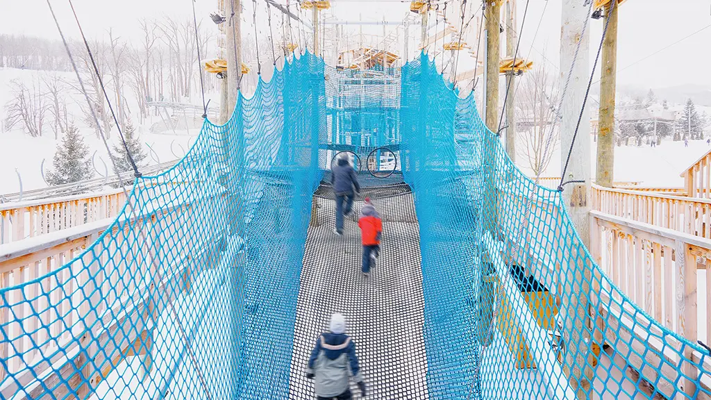  Tres niños caminan a lo largo de un circuito de cuerdas elevado de plataforma en plataforma con un paisaje invernal de fondo.