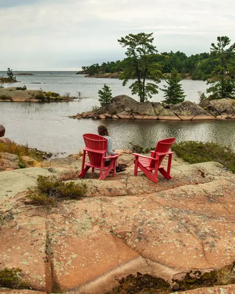 キラーニー近郊のジョージアン湾の海岸線沿いにある象徴的なピンク色の花崗岩の岩の上に置かれた 2 つの赤いマスコーカ チェアのうちの 1 つに座っている女性。