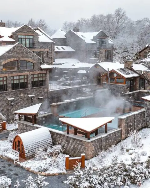 Des bâtiments historiques en pierre, légèrement saupoudrés de neige, entourent une piscine extérieure chauffée dans un complexe thermal.