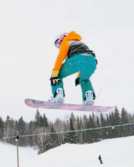 Un snowboarder en el aire realiza un salto en una pendiente nevada, con un bosque a lo lejos bajo un cielo nublado.