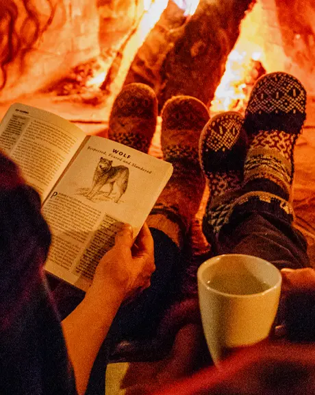 Dos personas se sientan una al lado de la otra frente a una chimenea encendida, con calcetines cómodos; una lee un libro mientras la otra sostiene una taza, creando una atmósfera invernal cálida e íntima.