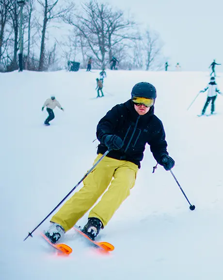 Un esquiador con pantalones amarillos desciende por una pendiente cubierta de nieve, mientras se ven otros esquiadores al fondo en un día de niebla.