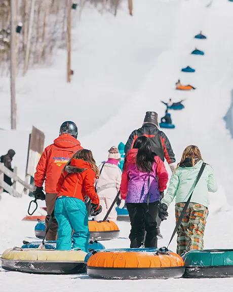 Families pull colorful snow tubes uphill at a tubing park, with snowy slopes and trees in the background.