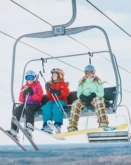 Tres personas viajan en un telesquí con tablas de snowboard y esquís, subiendo por una pendiente nevada en un claro día de invierno.