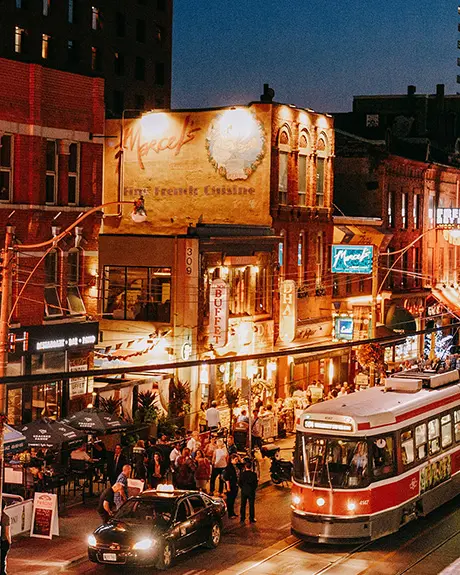 A crowded nightlife street with illuminated bars, restaurants, pedestrians and a streetcar passing through at night.
