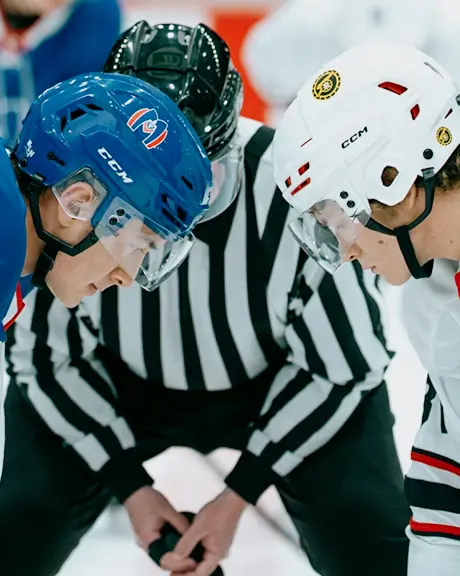 2 人のホッケー選手がパックを奪い合っており、1 人は青いジャージを着て、もう 1 人は白いジャージを着ています。審判が 2 人の間にしゃがんでいます。