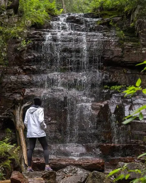 Ein Wanderer hält inne, um einen sanft plätschernden Wasserfall zu bewundern, der von moosbewachsenen Felsen, Bäumen und üppigem Grün im Norden Ontarios umgeben ist.