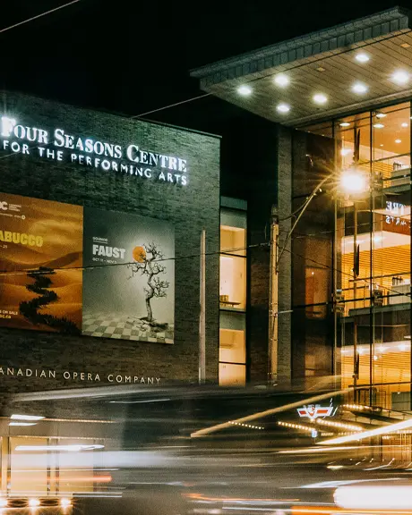 Edificio de artes escénicas con fachada de vidrio iluminado por la noche a lo largo de una concurrida calle de la ciudad.