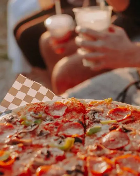 Une pizza fraîchement préparée, des gens tenant des boissons à proximité, sur la plage.