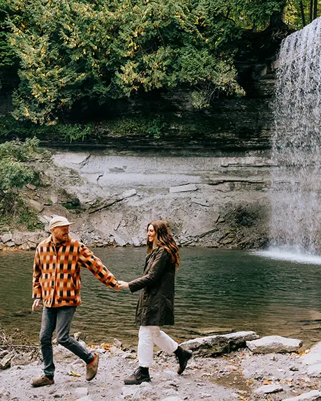 Couple walking hand in hand near a waterfall surrounded by forest.