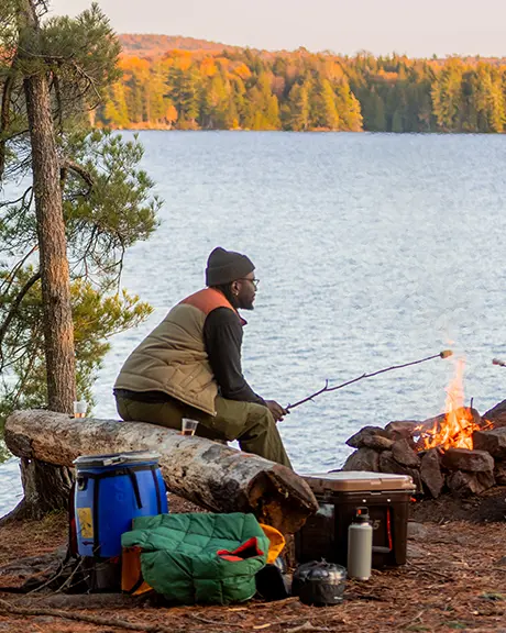  湖畔の岩場にある焚き火のそばに座った二人が、近くにキャンプ道具を置いてマシュマロを焼いている。