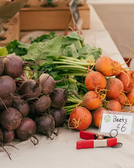  Frische Rüben und Gemüse werden auf einem Tisch auf einem Bauernmarkt präsentiert, im Hintergrund sind Blumen zu sehen.
