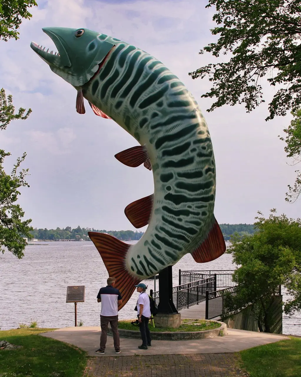 Dos visitantes se encuentran al pie de una gigantesca escultura al borde de la carretera que representa un pez lucio saltando, con marcas de rayas verdes y blancas y aletas de color óxido, montada sobre un poste alto a lo largo de la orilla del río.