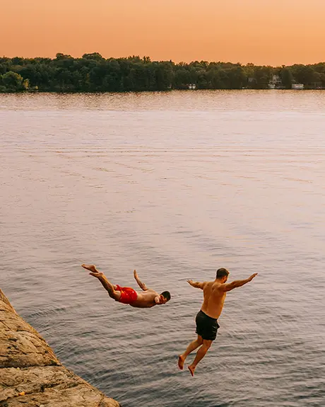 Two people jumping off a rocky cliff into a lake at sunset.