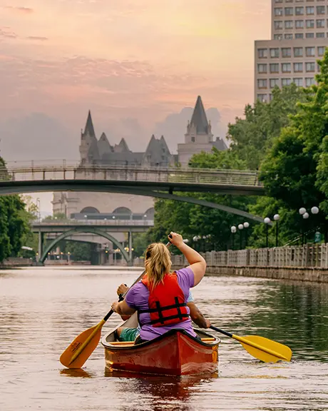 Zwei Personen paddeln bei Sonnenuntergang mit einem Kanu auf dem Rideau-Kanal in Ottawa; im Hintergrund sind Brücken und Stadtgebäude zu sehen.