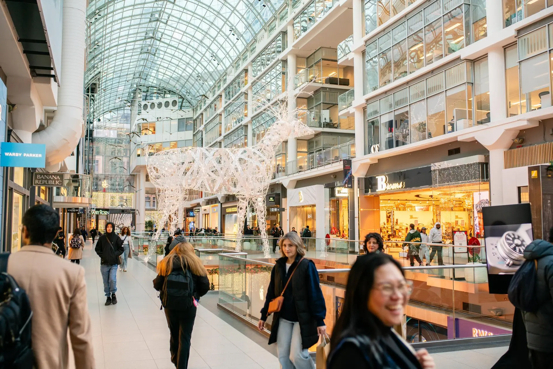 Shoppers fill Toronto's Eaton Centre mall during the holiday season, with large illuminated reindeer sculptures under the iconic vaulted glass ceiling.