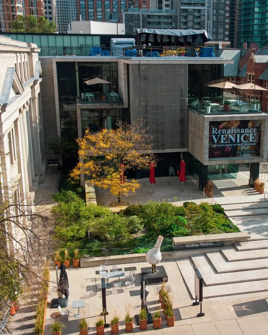 Aerial view of the Gardiner Museum in Toronto, featuring a modern building with rooftop terrace, landscaped courtyard, and outdoor sculptures, surrounded by the city skyline.