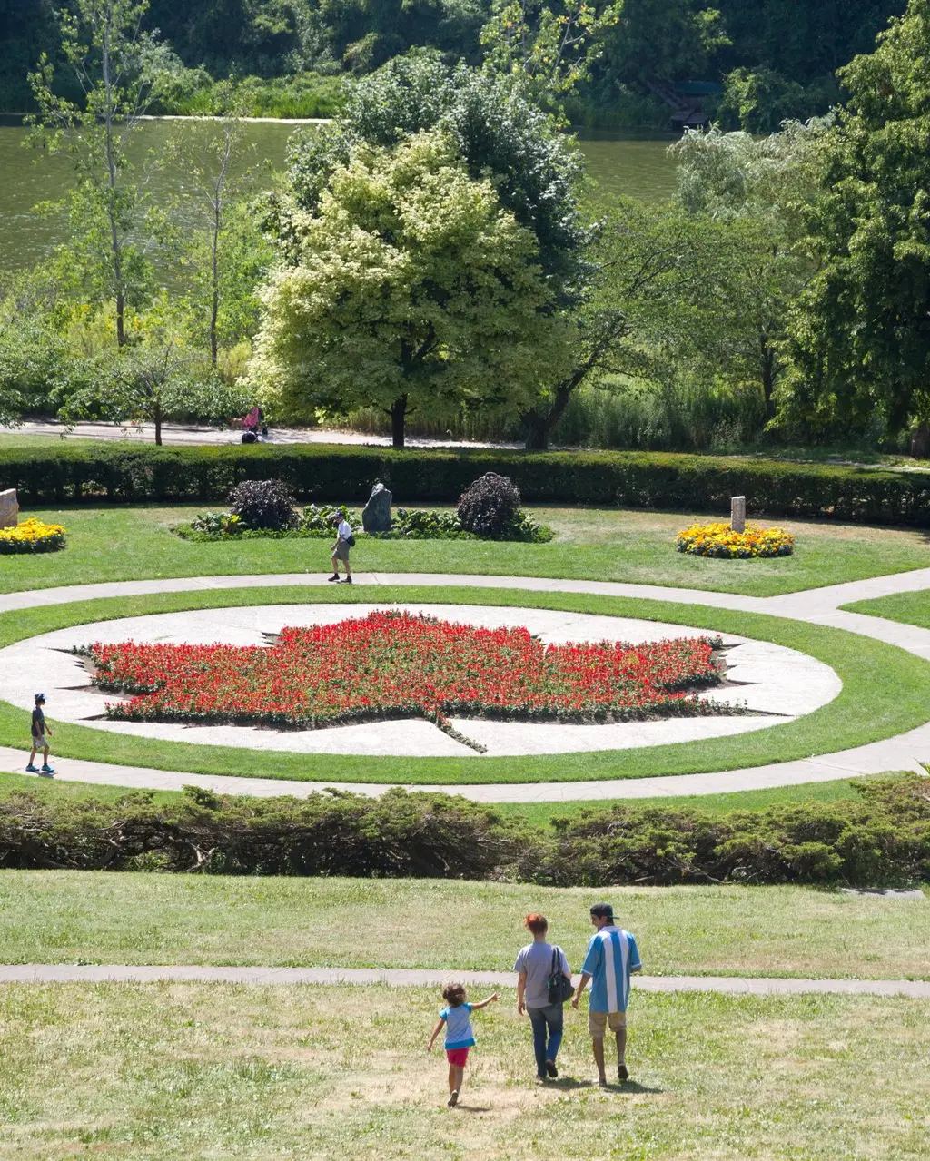 Aerial view of High Park's formal garden featuring a red maple leaf floral display, with a family walking toward it and a pond visible through the trees
