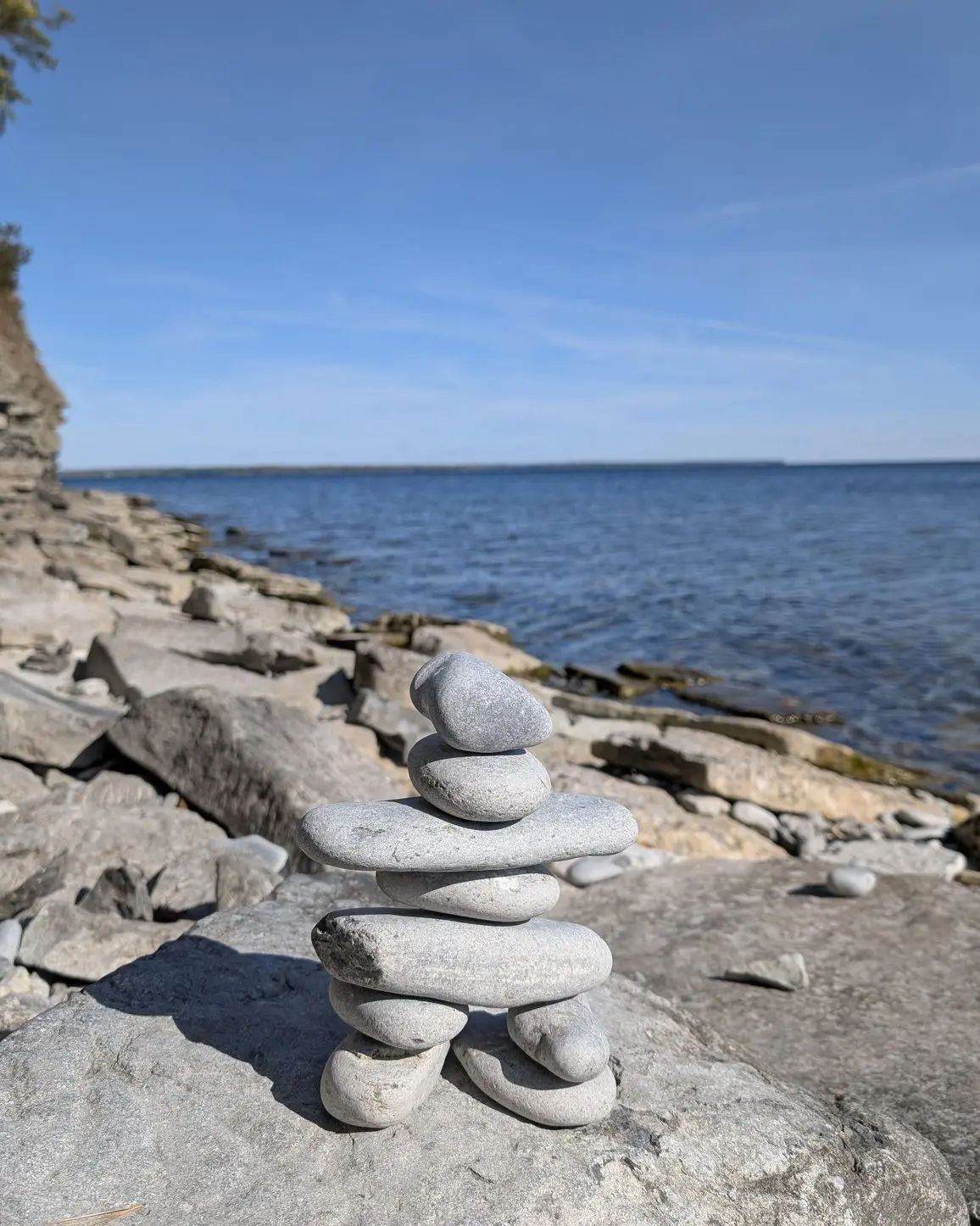 Stacked stone inukshuk on a rocky shoreline overlooking blue water and a limestone bluff.