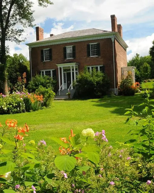 Casa histórica de ladrillo con porche ribeteado en blanco, rodeada de jardines y coloridas flores sobre un césped verde.