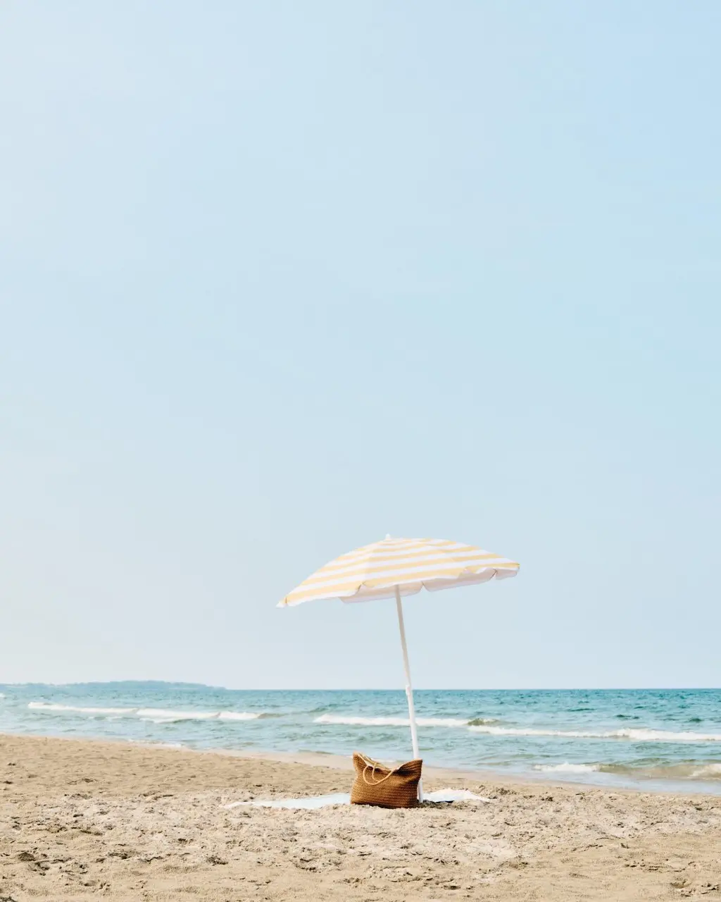 Sandy beach with a striped umbrella and tote bag by the water's edge.