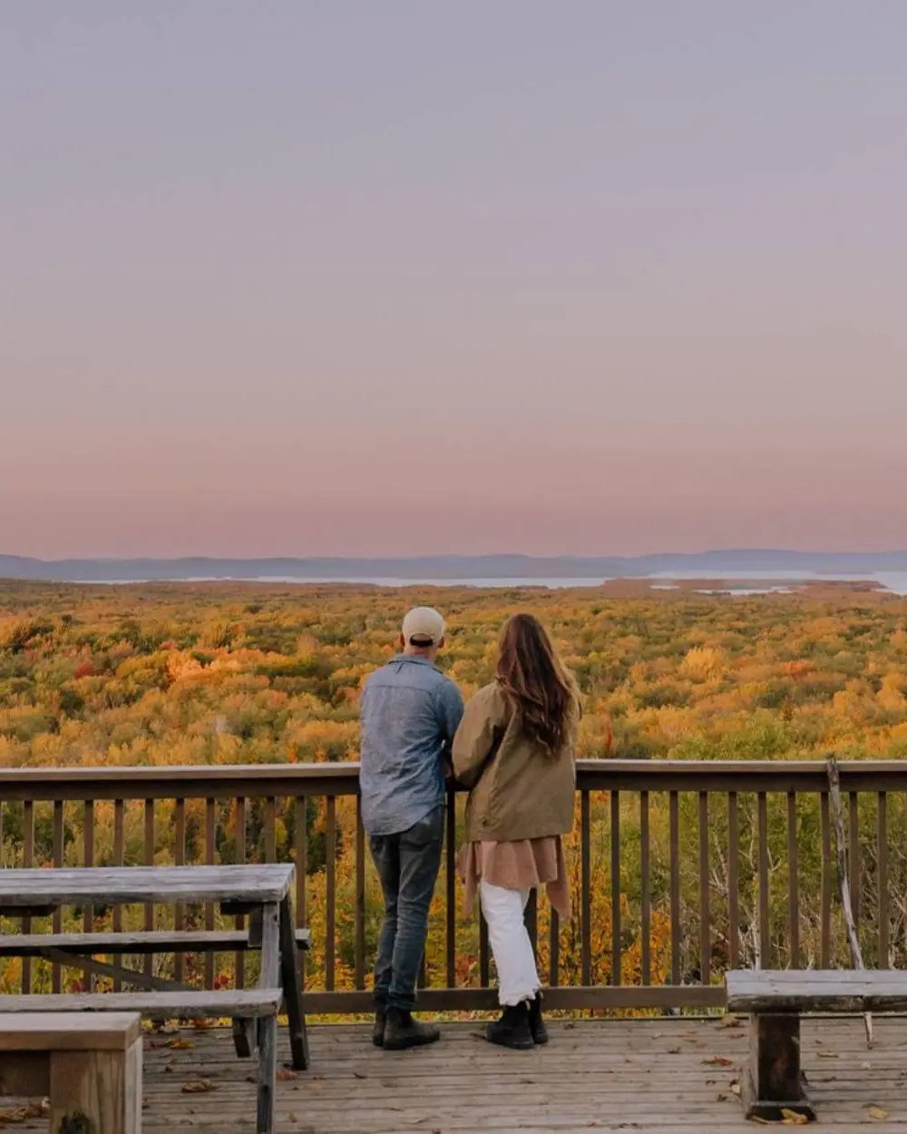 A couple standing on a wooden lookout platform admire the view of fall colour and a pink sky at dusk.