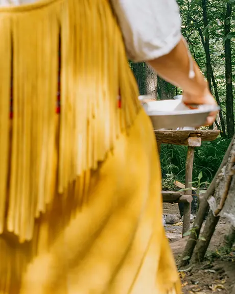 A woman carries a plate to a traditional Indigenous dwelling.