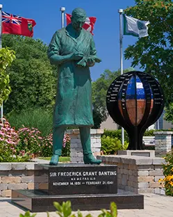 Statue en bronze de Sir Frederick Grant Banting sur un socle en pierre, avec des drapeaux et une sculpture de globe dans le jardin situé derrière.