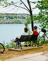 Dos personas dejan sus bicicletas a un lado de un sendero, sentadas en un banco negro, contemplando un lago con árboles al fondo.