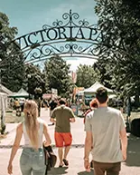Menschen gehen unter einem kunstvoll verzierten Eisenbogen mit der Aufschrift „Victoria Park“ hindurch in Richtung eines Festivals mit Zelten und Verkaufsständen.