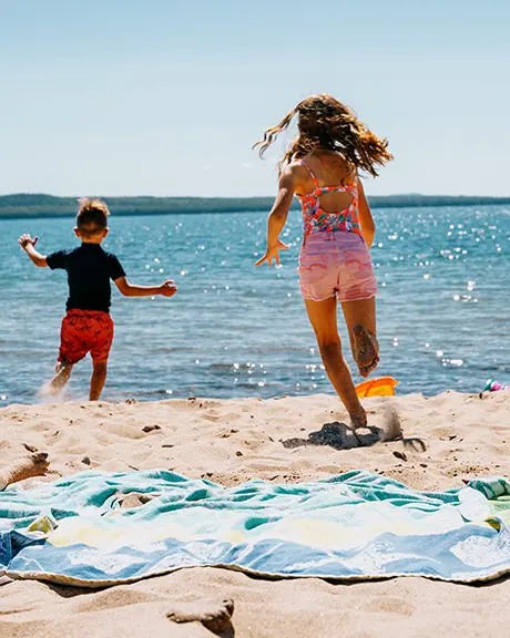 Two adults relax on towels at a sandy lakeside beach while two children run toward the water on a sunny day with clear blue sky.