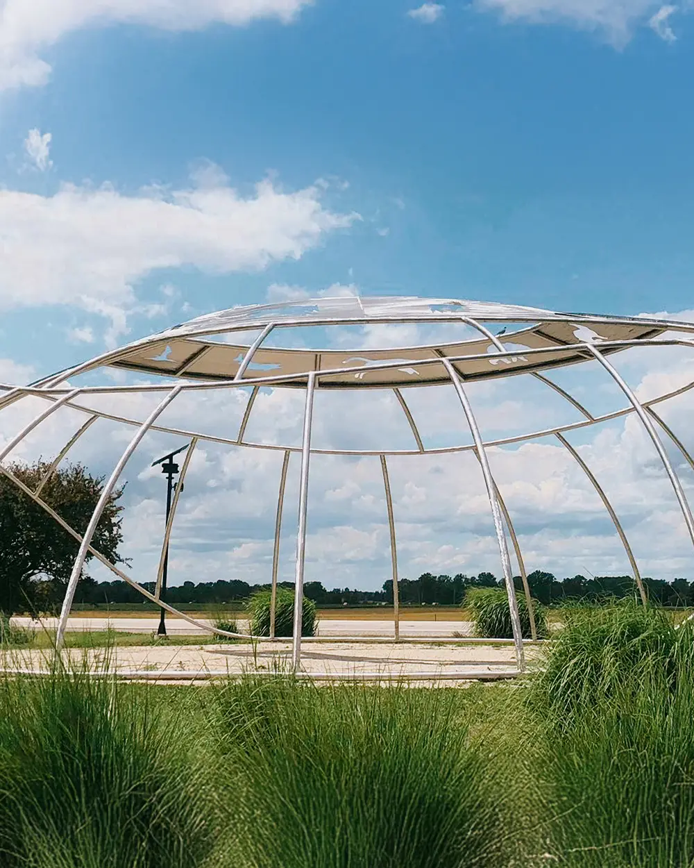Chatham-Kent Structure métallique sculpturale en plein air avec une armature géométrique courbée, entourée de hautes herbes, avec des terres agricoles et un ciel bleu partiellement nuageux en arrière-plan.