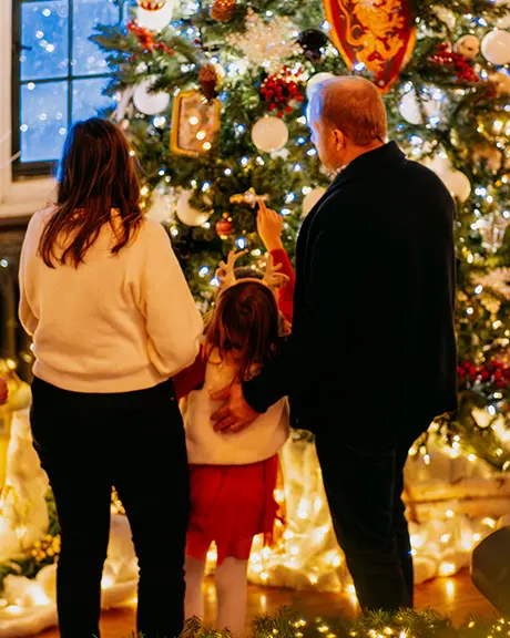 Family admiring a decorated Christmas tree surrounded by festive lights and ornaments indoors.
