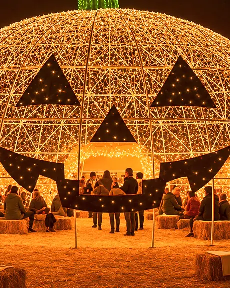 Une grande structure illuminée en forme de citrouille, décorée d’un visage de citrouille-lanterne, entourée de bottes de foin et de personnes lors d’un événement d’Halloween en plein air. 