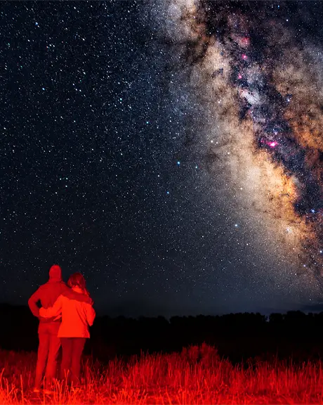 Deux personnes se tiennent côte à côte dans un champ herbeux illuminé par une lumière rouge, contemplant le ciel nocturne étoilé et la Voie lactée lumineuse.
